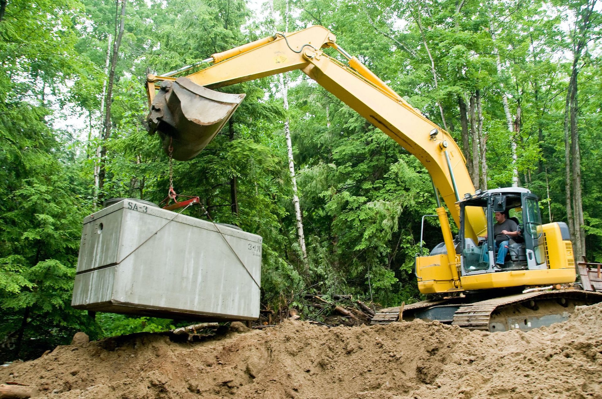 Yellow excavator lifting a large concrete structure in a forested area.